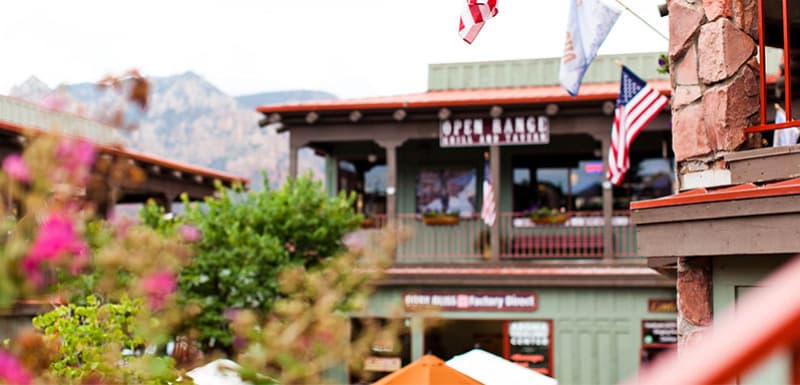 На изображении показано здание с деревянным балконом и вывеской «OPEN RANGE GRILL & TAVERN». На балконе развеваются американские флаги. На переднем плане — зелёные и ярко-розовые цветы, а также растения, частично размытые.